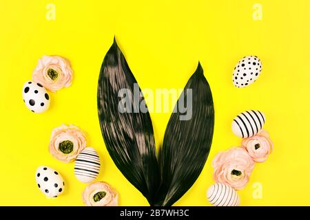 Bunny ears made of natural green leaves with decorative eggs and ranunculus flowers on yellow background. Easter concept. Stock Photo