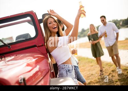 Happy young women drinks cider from the bottle by the convertible car with friends Stock Photo