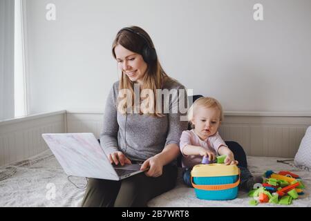 Young Caucasian mother blogger with baby working on laptop from home. Workplace of freelance woman student with kid toddler. Stay at home single mom e Stock Photo
