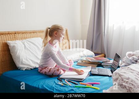 Caucasian girl child sitting in bed and learning online on laptop Internet. Virtual class lesson on video during self isolation at home. Distant remot Stock Photo