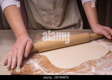 A woman rolls out the dough. Plywood cutting board, wooden flour sieve and wooden rolling pin - tools for making dough Stock Photo