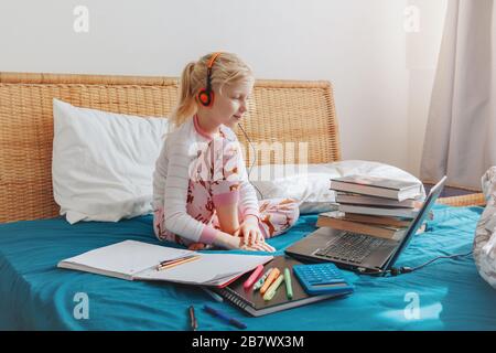 Caucasian girl child sitting in bed and learning online on laptop Internet. Virtual class lesson on video during self isolation at home. Distant remot Stock Photo