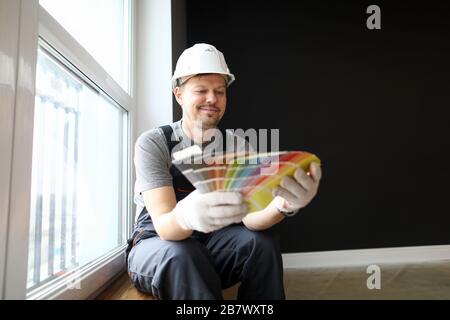 Young repairman in helmet sitting in flat and choosing color for interior Stock Photo