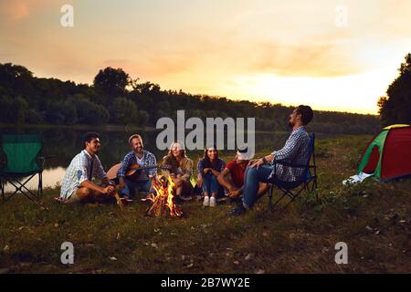 A group of people sitting by the bonfire next to the tent at night in the summer in autumn. Stock Photo