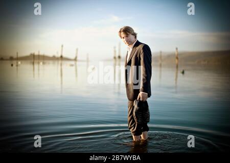 Portrait of a mid-adult man wearing a suit while standing in shallow water with his pants rolled up at a beach. Stock Photo