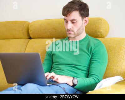 Young caucasian man working from home on his computer sitting on a sofa, since the coronavirus outbreak in Europe. Stock Photo