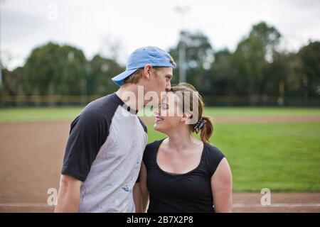 Teenage boy kissing his girlfriend on the forehead next to a sports field. Stock Photo