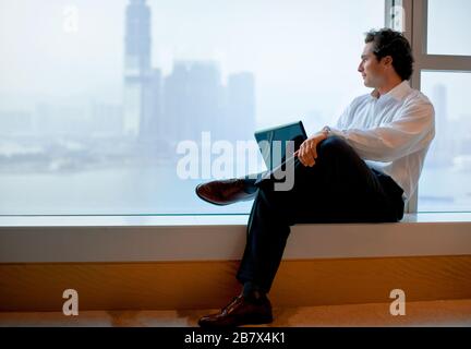 Businessman using his laptop, sitting on a window ledge looking out over the city skyline. Stock Photo