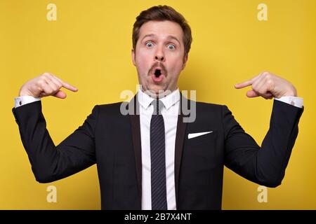 Positive young man with mustache smiling and pointing at himself with index fingers. Handsome man wearing suit gesturing and looking at camera. Self p Stock Photo
