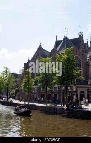 View of people riding a small boat on canal doing a cruise tour in front of 13th-century The Oude Church in Amsterdam. Many people hang out on street. Stock Photo