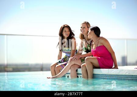 Group of smiling friends relaxing on the edge of a swimming pool. Stock Photo