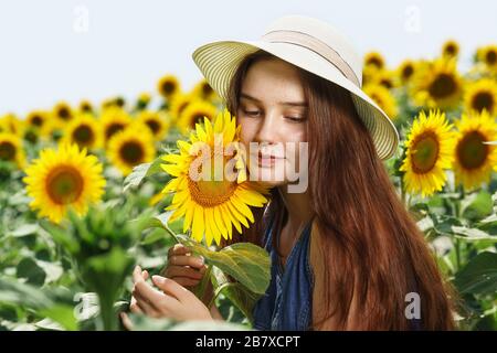 A beautiful teenage girl in a denim dress and a hat with a brim stands in a field with sunflowers. Portrait. Close up Stock Photo