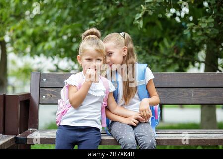 Two small children tell secrets on a bench. Girls with backpacks. The concept is back to school, family, friendship and childhood. Stock Photo