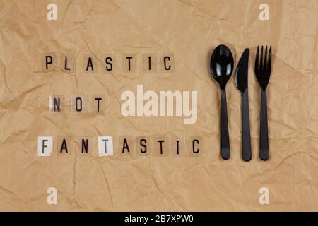Disposable black appliances fork, spoon, knife, phrase from letters on transparent base Plastic not fantastic on brown crumpled craft paper. Eco, zero Stock Photo