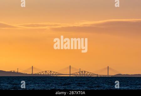 Firth of Forth, Scotland, United Kingdom. 18th March 2020. UK Weather: a colourful orange sky at sunset over the distinctive outline of the three Forth bridges seen from East Lothian Stock Photo