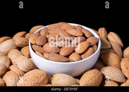 Peeled almonds in a white bowl surrounded by raw almonds in shell. Isolated on black background. Stock Photo