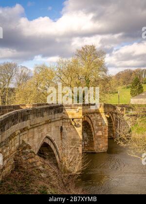 An ancient arched bridge across the River Derwent.  Trees are nearby and there are fields in the background. A cloudy sky is above. Stock Photo