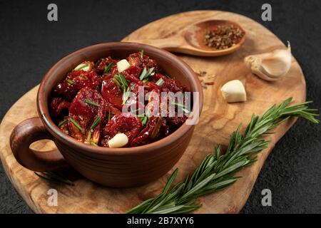 Sun-dried tomatoes with garlic, rosemary and spices in a clay bowl on an olive wood cutting board. Stock Photo