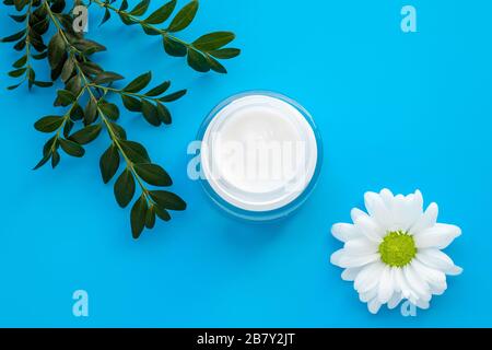 Face cream jar with white chamomile flower and green branch on a blue background. Herbal lotion in a glass container, natural cosmetic. Skin care in d Stock Photo