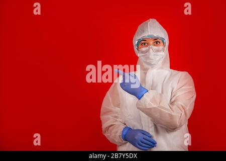 A woman in a protective suit against the COVID-19 virus indicates the direction. Medic woman wearing protective clothing against coronovirus on a red background. Stock Photo
