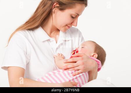 Unidentified young mother holds in her arms a charming sleeping little girl with a pacifier in her mouth on a white background. Concept of caring for Stock Photo