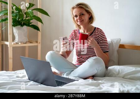 Distance learning online education and work.Happy woman girl working office work remotely from home on bed. Using computer and drinking coffee Stock Photo
