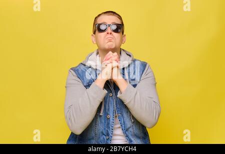 Stylish Caucasian man in a jeans and sunglasses on a yellow background. Close up. Crossing his hands plaintively looks at the top. Stock Photo