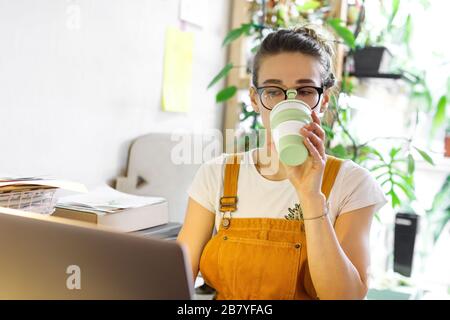 Young female gardener in glasses using laptop, communicates on internet with customer in home garden/greenhouse, drinking coffee/tea from reusable mug Stock Photo