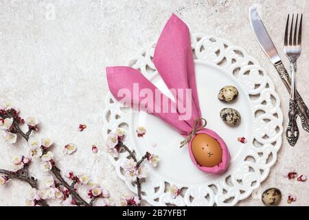 Beautiful festive Easter table setting with egg in napkin like Easter Bunny and blossoming branches on light textured background. Top view with copy s Stock Photo