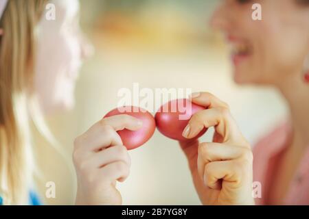 Closeup on smiling mother and daughter having egg tapping with red easter eggs at home in sunny spring day. Stock Photo