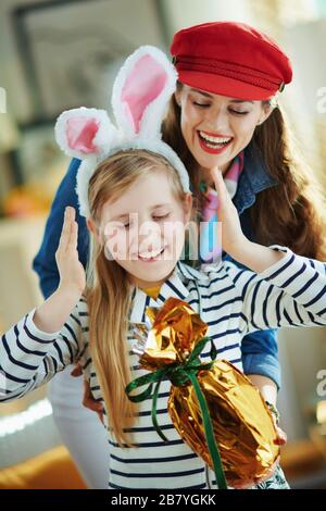 mother presenting wrapped in gold foil big easter egg to daughter in easter bunny ears in the modern house in sunny spring day. Stock Photo
