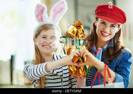 Portrait of happy trendy mother and child with wrapped in gold foil big easter egg and shopping bags showing heart shaped hands at modern home in sunn Stock Photo