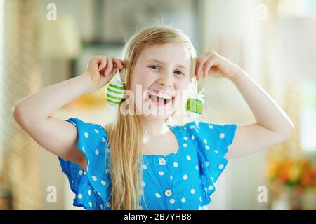 happy modern child in a polka dot blue overall and easter bunny ears making earrings with green decorative easter eggs in the modern house in sunny sp Stock Photo