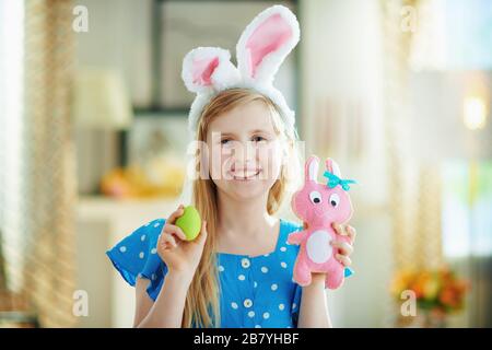 happy modern girl in a polka dot blue overall and easter bunny ears at modern home in sunny spring day showing pink rabbit toy and green easter egg. Stock Photo