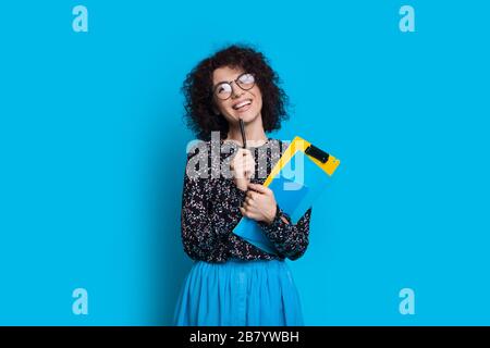 Charming curly haired student holding some books while posing in a dress on blue wall with blank space Stock Photo
