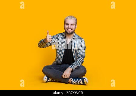 Blonde caucasian man with beard gesturing the like sign while sitting on a yellow wall Stock Photo