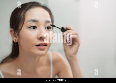 Young woman dyes her eyelashes with mascara brush. Young beautiful woman applying mascara makeup on eyes at bathroom. Stock Photo