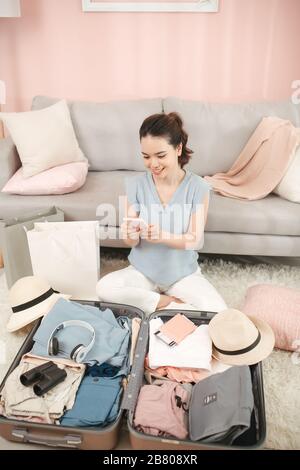 Upper view of smiling elegant woman at modern home in sunny summer day using mobile travel agent app on smartphone near open travel suitcase. Stock Photo