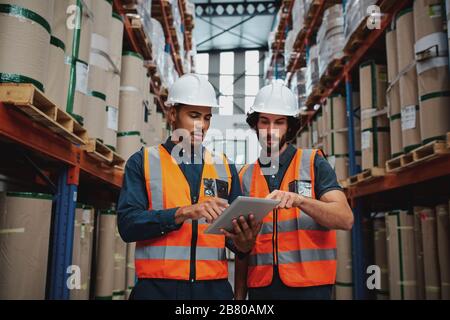Focused manager and employee standing in warehouse discussing inventory stock while standing between cardboard packed goods with safety hardhat and Stock Photo