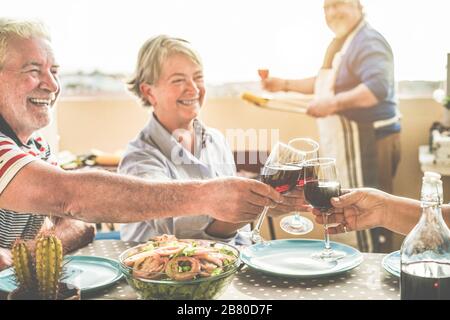 Senior friends cheering with red wine at barbecue dinner in patio outdoor - Mature people dining and laughing togheter - Joyful elderly lifestyle and Stock Photo