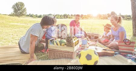 Happy families doing picnic in nature park outdoor - Young parents having fun with children in summer eating fresh fruit and laughing together - Love