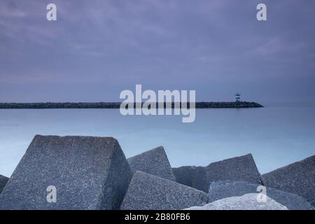 Large blocks of grey stone with a lighthouse in the distance, at dusk, Scheveningen, the Netherlands Stock Photo