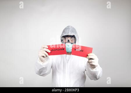 Man in surgical mask wearing white protective suit and a red sign with the word coronavirus split in half isolated in studio on white background. Prev Stock Photo
