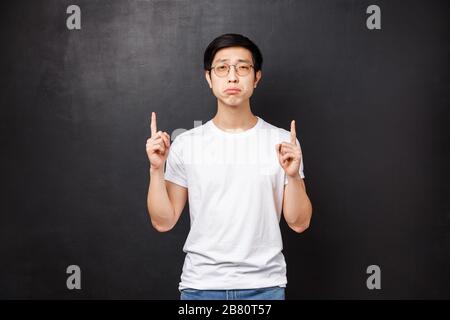 Portrait of gloomy and upset distressed young asian guy whining, complaining on unfair life, pointing fingers up with regret and jealous, look camera Stock Photo