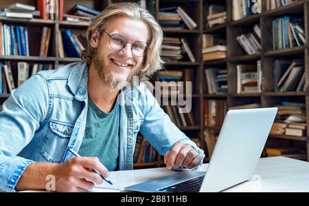 Smiling male student study online on computer make notes in library, portrait. Stock Photo