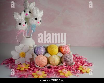 3 Easter bunnies in a white vase with a ceramic egg holder containing 6 Easter eggs on top of a bed of pink paper shreds with felt daisy flowers.  Eas Stock Photo