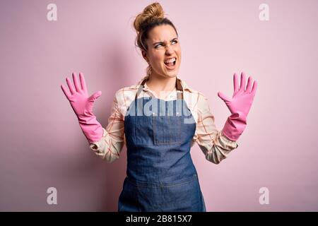 Young beautiful blonde cleaner woman doing housework wearing arpon and gloves crazy and mad shouting and yelling with aggressive expression and arms r Stock Photo