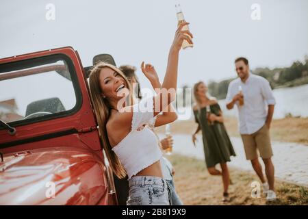 Happy young women drinks cider from the bottle by the convertible car with friends Stock Photo