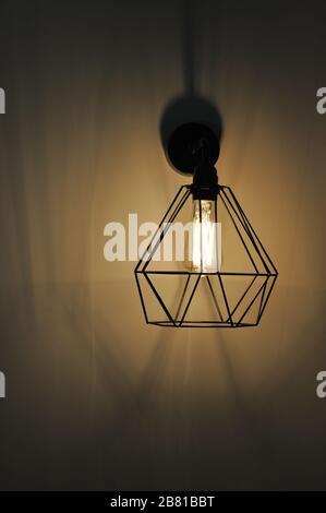 Modern pendant light made of diamond shape wire carcass with glass bulb inside. Glowing filament in lamp of minimal style. Sconce lamp hanging on wall Stock Photo