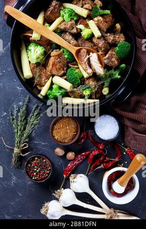 Black dish with roasted pork pieces, broccoli, corn, onion and garlic and  ingredients on a dark concrete background, flat lay, vertical orientation Stock Photo